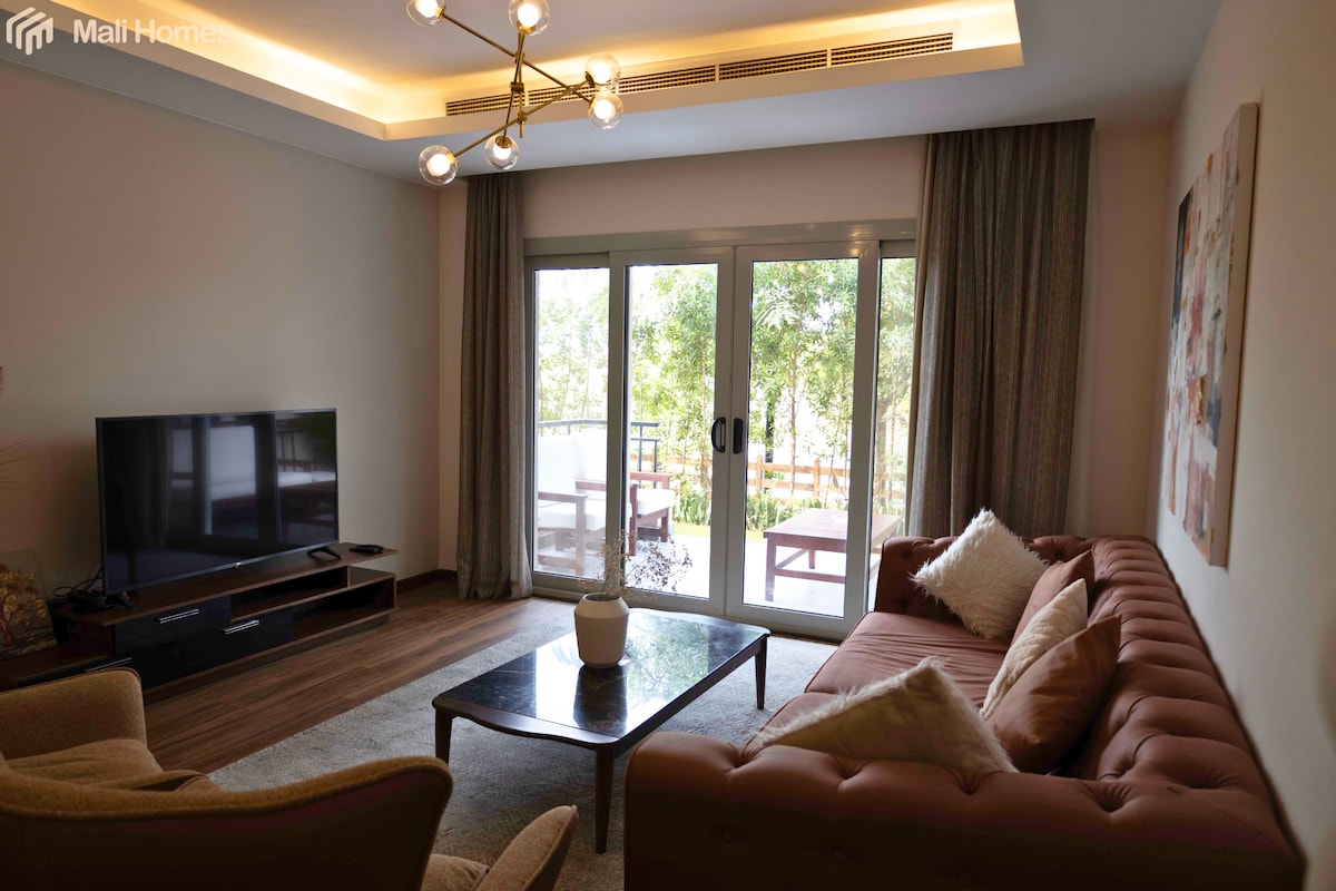 A comfortable living room is displayed, featuring a plush brown sofa and a glass coffee table. Large sliding doors provide access to a private outdoor space, while a television is positioned on a cabinet. Soft lighting enhances the gentle palette of neutral tones.