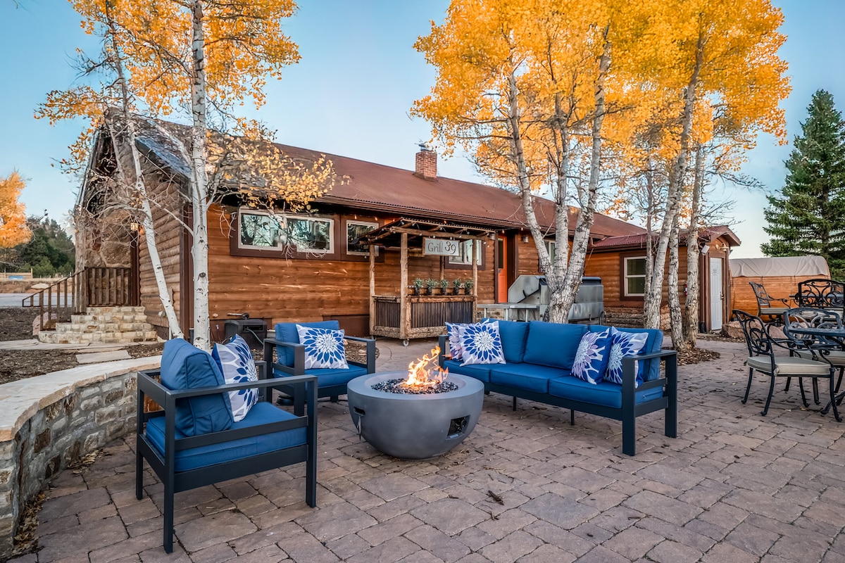 An outdoor patio area features a circular stone fire pit surrounded by deep blue seating. Nearby, trees with golden leaves stand tall against a rustic wooden cabin backdrop, creating a warm and inviting atmosphere. Tables and chairs are arranged for communal dining in the background.