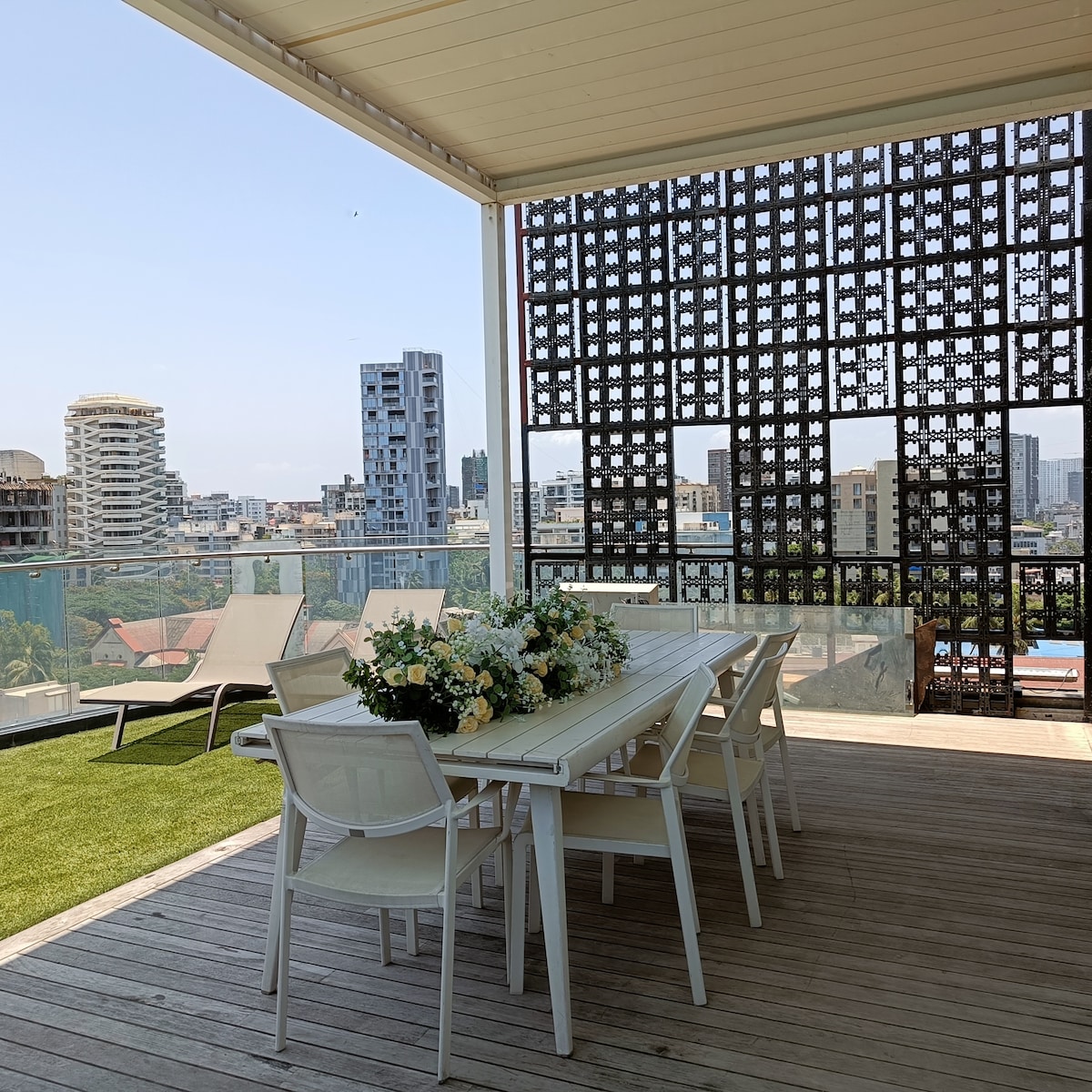 An outdoor dining area is presented with a white wooden table surrounded by four chairs. A bouquet of flowers is centered on the table. Behind, a view of the city skyline is visible, along with lounge chairs positioned for relaxation.