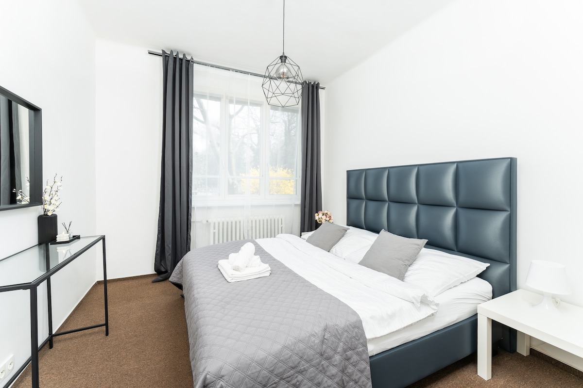 A well-furnished bedroom features a spacious bed with a padded headboard. Soft linens in neutral tones are layered with decorative pillows. A large window allows natural light to fill the room, complemented by dark curtains. A simple bedside table and a minimalist desk complete the space.