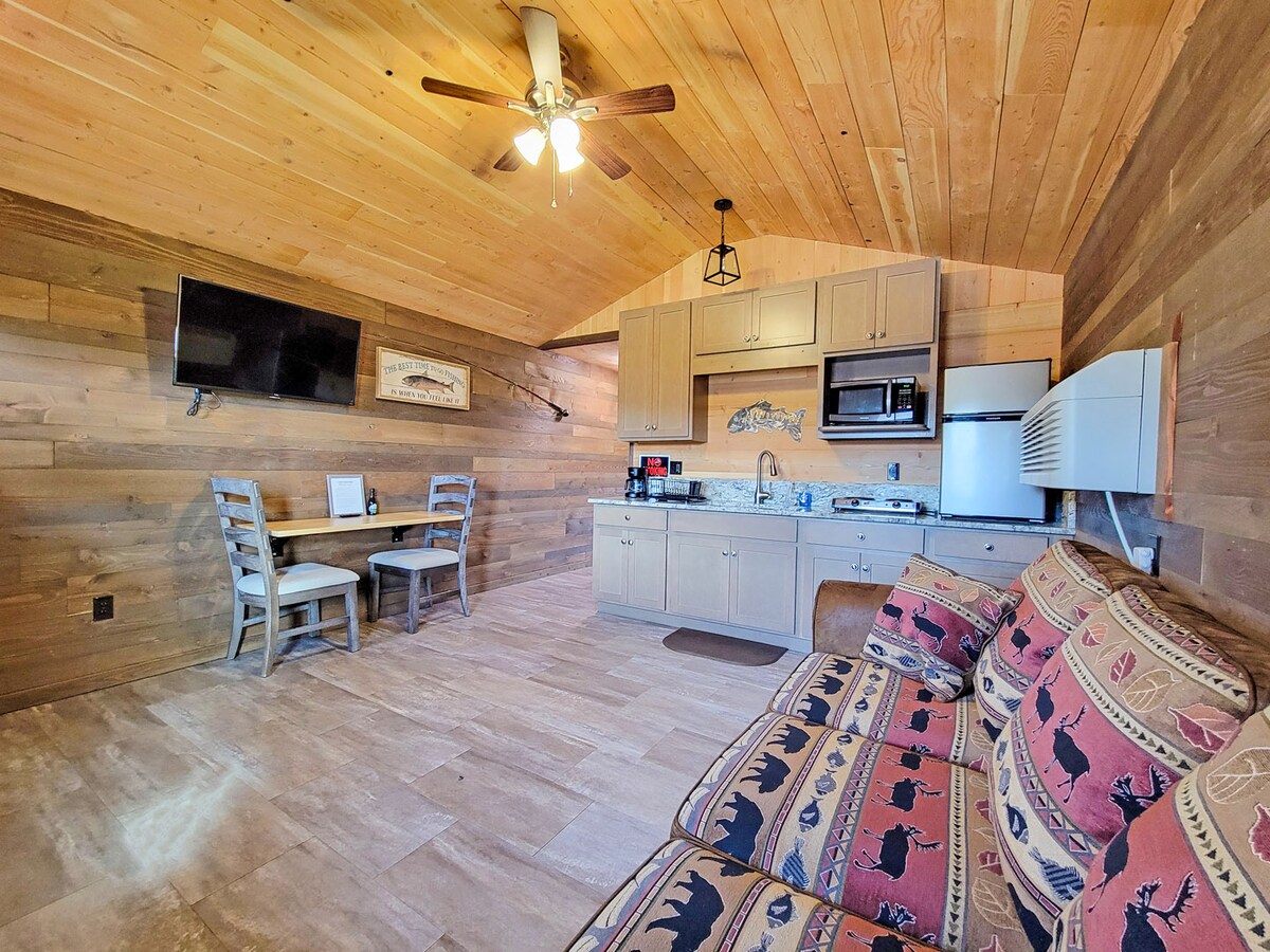 A cabin interior features wooden walls and a high ceiling, creating a rustic atmosphere. A kitchenette is equipped with a microwave, refrigerator, and stove. A small dining area is set up beside a flat-screen TV. Comfortable seating with patterned cushions is arranged for relaxation.