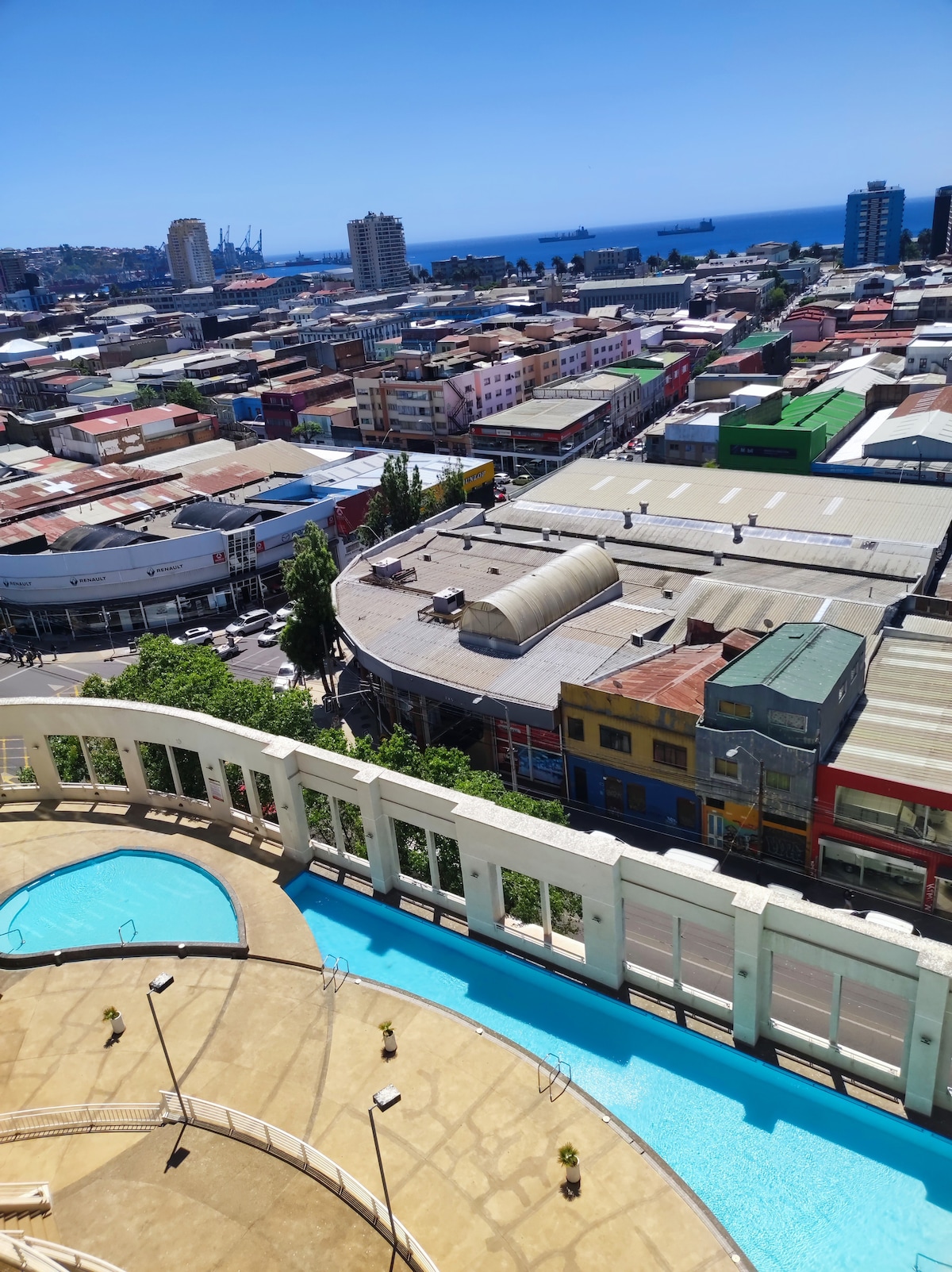 A panoramic view captures the bustling cityscape featuring colorful buildings and rooftops. In the foreground, a sparkling pool is visible, framed by a sleek stone perimeter. The distant ocean is glimpsed, complementing the vibrant urban scene below.