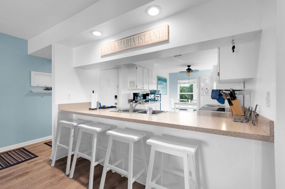 A bright kitchen space is highlighted by a spacious countertop with four high-backed white stools. Light blue walls create a refreshing backdrop, while a sign reading 'ANNA MARIA ISLAND' adorns the wall. Natural light flows in from windows, enhancing the airy feel of the area.