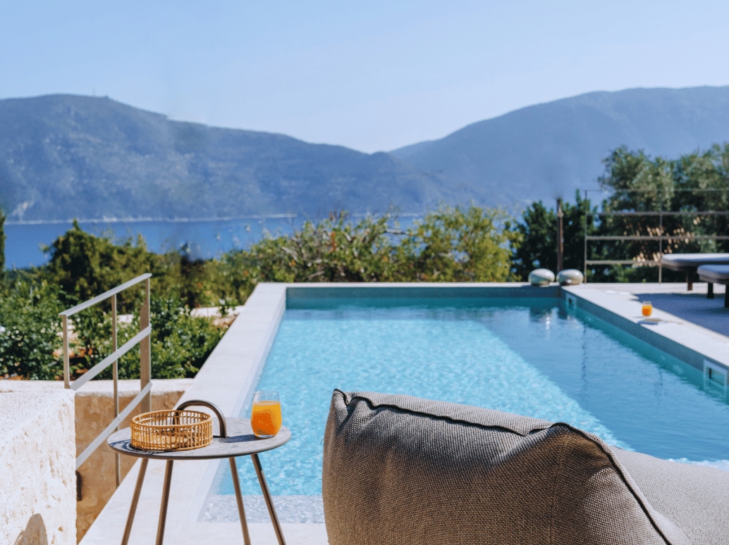 A crystal-clear swimming pool glistens under the sun, offering views of rolling hills and the sea in the distance. A cozy lounge chair and a side table, holding a refreshing drink, provide a comfortable spot to relax. Lush greenery surrounds the space, enhancing the serene atmosphere.