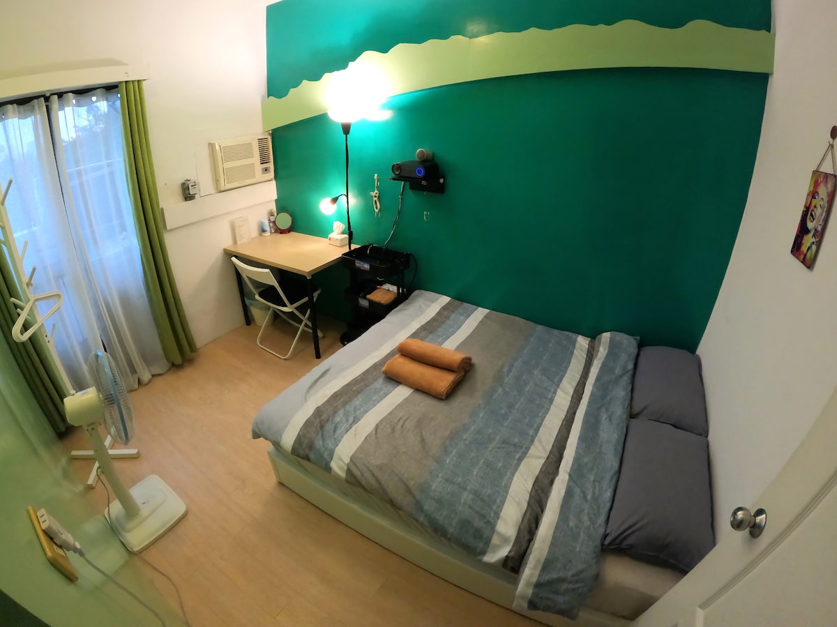 A cozy sleeping area features a spacious bed with a striped blanket and towels neatly arranged on top. A study desk is positioned against the vibrant green wall, accompanied by a small lamp and a fan. Floor-to-ceiling curtains allow for natural light to filter in.