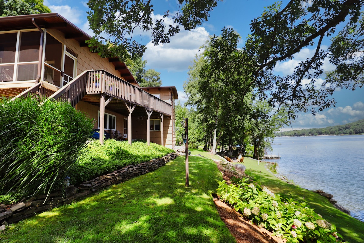 A lakeside view of the home reveals a spacious deck leading to lush green lawns and colorful shrubs. The tranquil waters of Candlewood Lake frame the background, with trees providing natural shade and a serene atmosphere. A path leads directly to the water's edge.