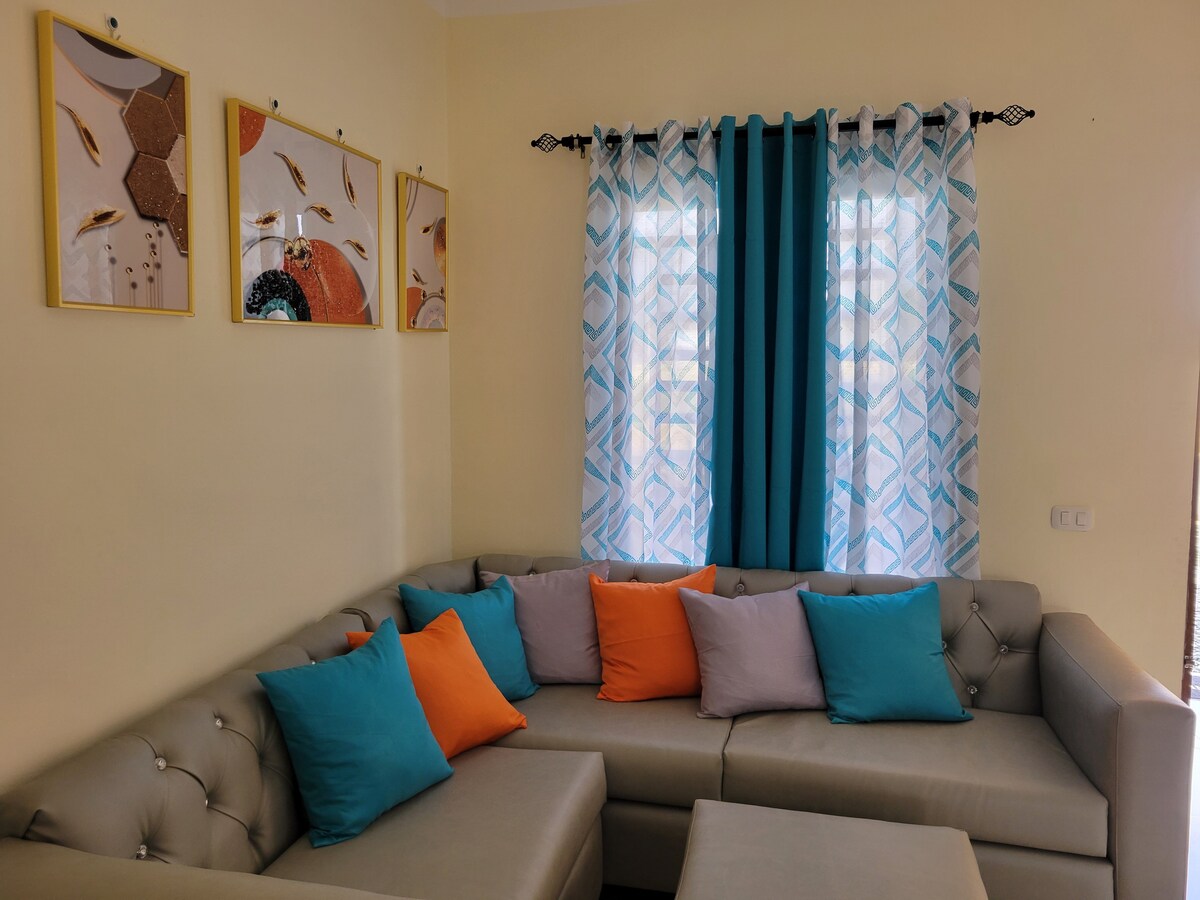 A spacious living area features a comfortable sectional sofa adorned with vibrant cushions in shades of turquoise and orange. Colorful artwork complements the light yellow walls, while sheer curtains allow natural light to fill the room.
