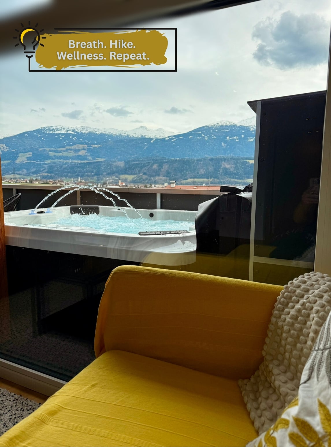 A cozy seating area features a yellow couch with a textured throw pillow. Through large windows, a private whirlpool is visible, with mountain views showcasing snow-capped peaks and expansive sky. Natural light enhances the serene atmosphere, inviting relaxation and enjoyment of the scenic landscape.