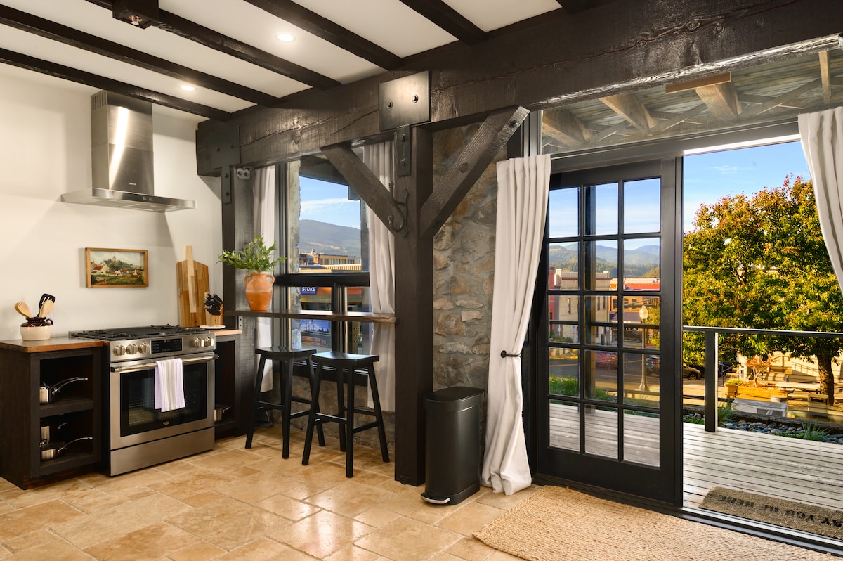 A modern kitchen is seen, featuring stainless steel appliances, including a stove and range hood. A wooden bar table with two stools sits by large glass doors that open to a balcony, offering views of the surrounding landscape and vibrant outdoors.