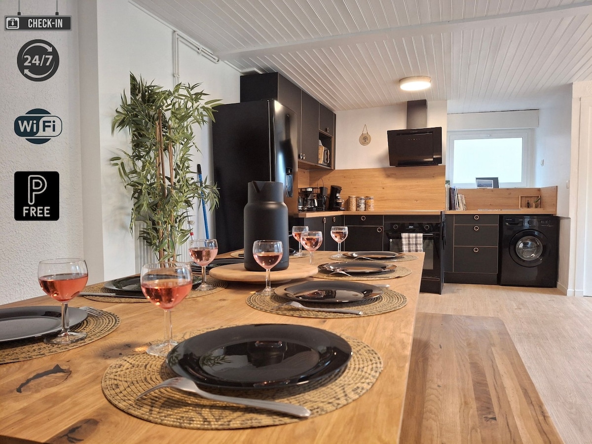 A spacious dining area is highlighted by a wooden table set for six, featuring black plates and glasses filled with pink beverages. The adjacent kitchen showcases modern appliances, including a sleek black refrigerator and a washing machine, surrounded by warm wooden accents.