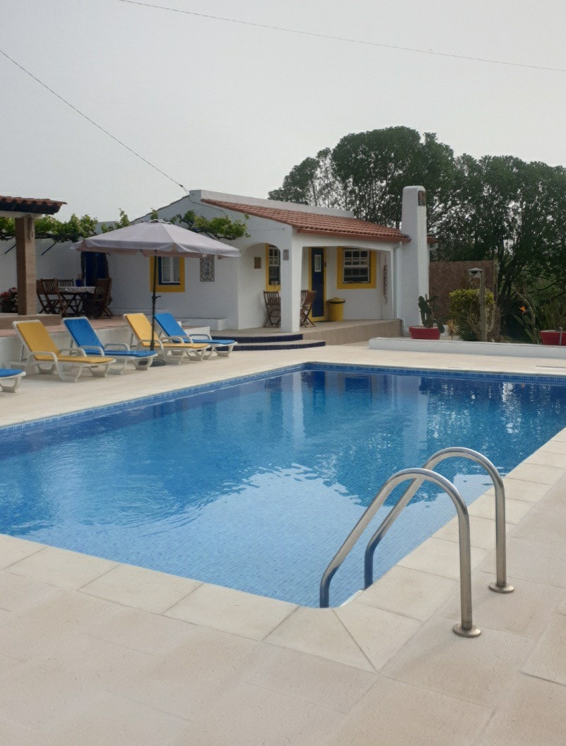A clear swimming pool is framed by a sun terrace, complete with several lounge chairs. An umbrella provides shade for the seating area, while a cottage with yellow accents stands in the background, surrounded by greenery. The scene offers a peaceful outdoor environment.
