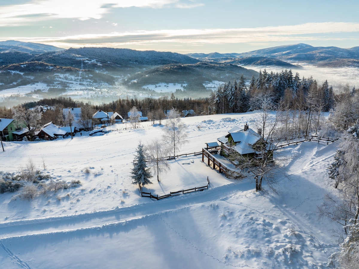 A panoramic view of the winter landscape showcases a cottage surrounded by snow-covered fields and trees. The scene features distant hills blanketed in snow under a clear blue sky, creating a serene atmosphere that highlights the beauty of the natural surroundings.