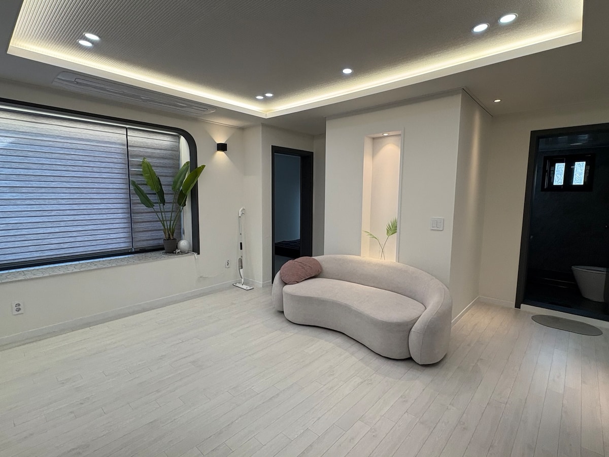An open living space features a modern, curved sofa in a soft texture, complemented by a small decorative pillow. Natural light filters through a large window, framed by greenery. Soft lighting enhances the minimalist design, creating a peaceful environment.