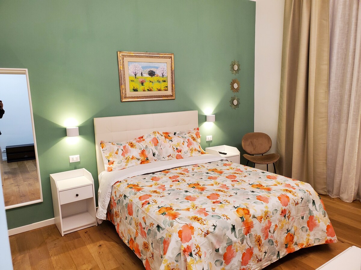 A bright bedroom features a spacious bed adorned with a floral-patterned cover. Soft green walls complement the room, while bedside tables and a chair provide functional furnishings. Natural light enters through curtains, enhancing the inviting atmosphere.