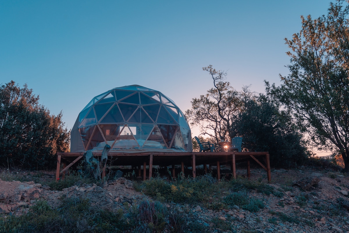 A geodesic dome is situated on a wooden deck, surrounded by lush greenery. The dome's transparent structure allows for unobstructed views of the sky and landscape, with a serene sunset illuminating the scene. Outdoor seating can be seen on the deck.