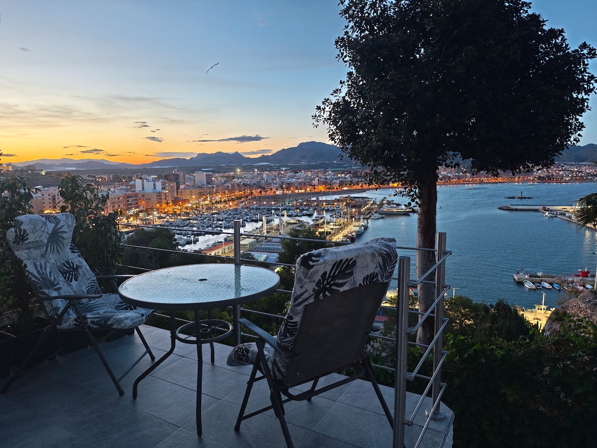 An outdoor terrace features a glass table and two chairs with floral patterns, surrounded by lush greenery. The captivating view showcases the marina and town illuminated at sunset, with mountains in the background and soft hues of orange and pink in the sky.