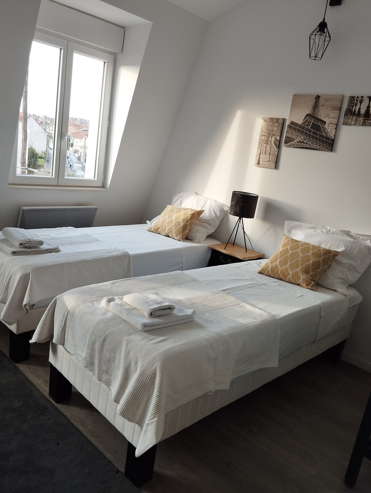 A bright sleeping area features two single beds, each dressed in crisp white linens. Decorative pillows and a nightstand with a lamp are present. Natural light streams in through a large window, enhancing the serene ambiance of the room.
