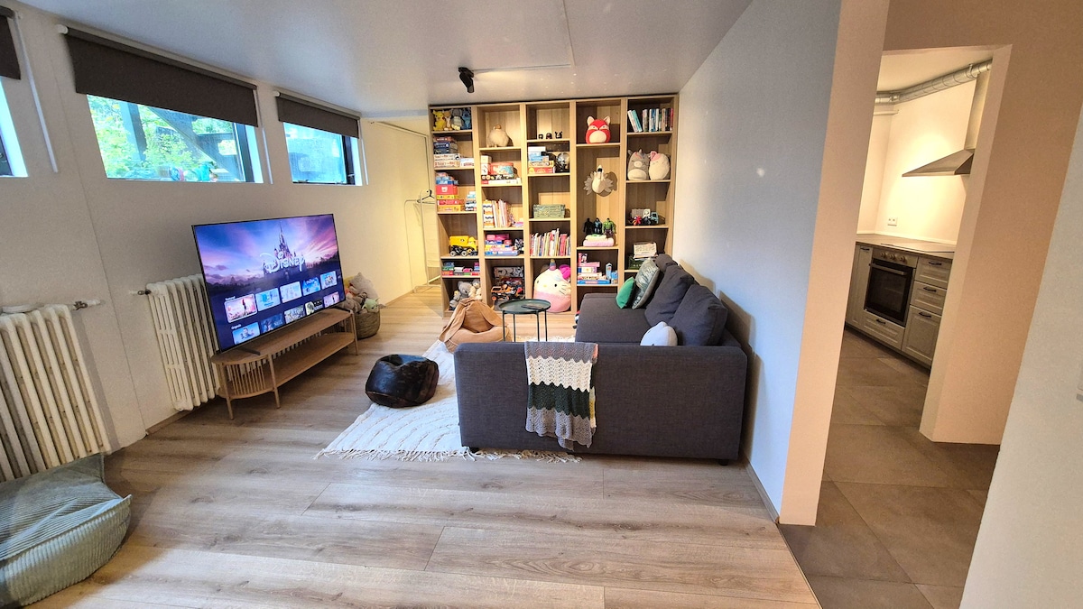 A welcoming living room is presented, featuring a spacious grey sofa and a large TV on a wooden stand. A well-organized shelf displays various toys. Light enters through multiple windows, illuminating the warm wood floors that extend into an adjoining kitchen area.