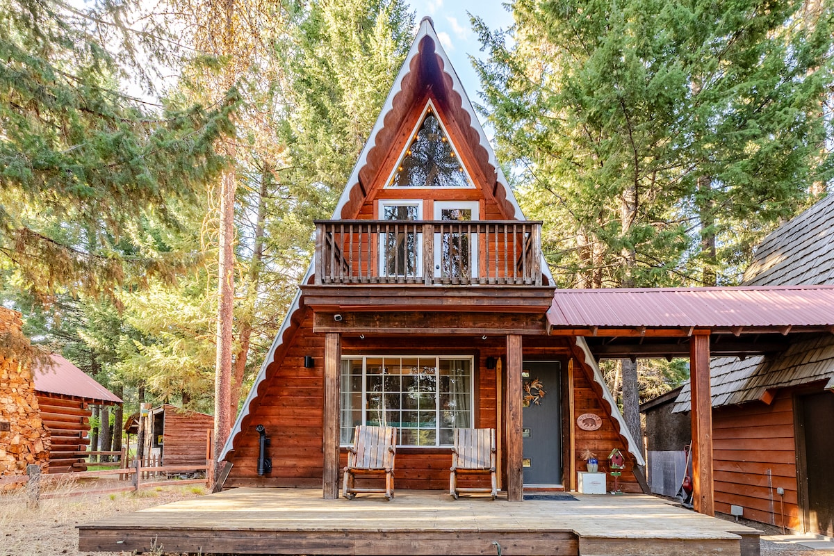 A charming cedar A-frame cabin is nestled among tall evergreens. The wooden porch features two rustic rocking chairs, inviting relaxation. A balcony above has a classic A-frame roofline, while large windows offer a view of the surrounding nature.