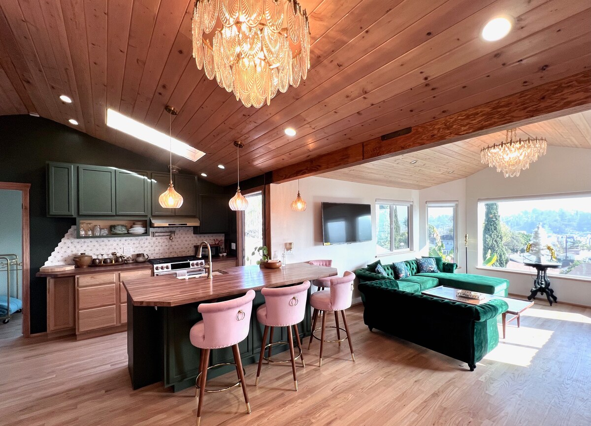 An open concept living area features a modern kitchen with a large island and bar stools. Elegant light fixtures hang from the wooden ceiling. Large windows offer a view of the surrounding hills, while plush seating adds comfort to the space.