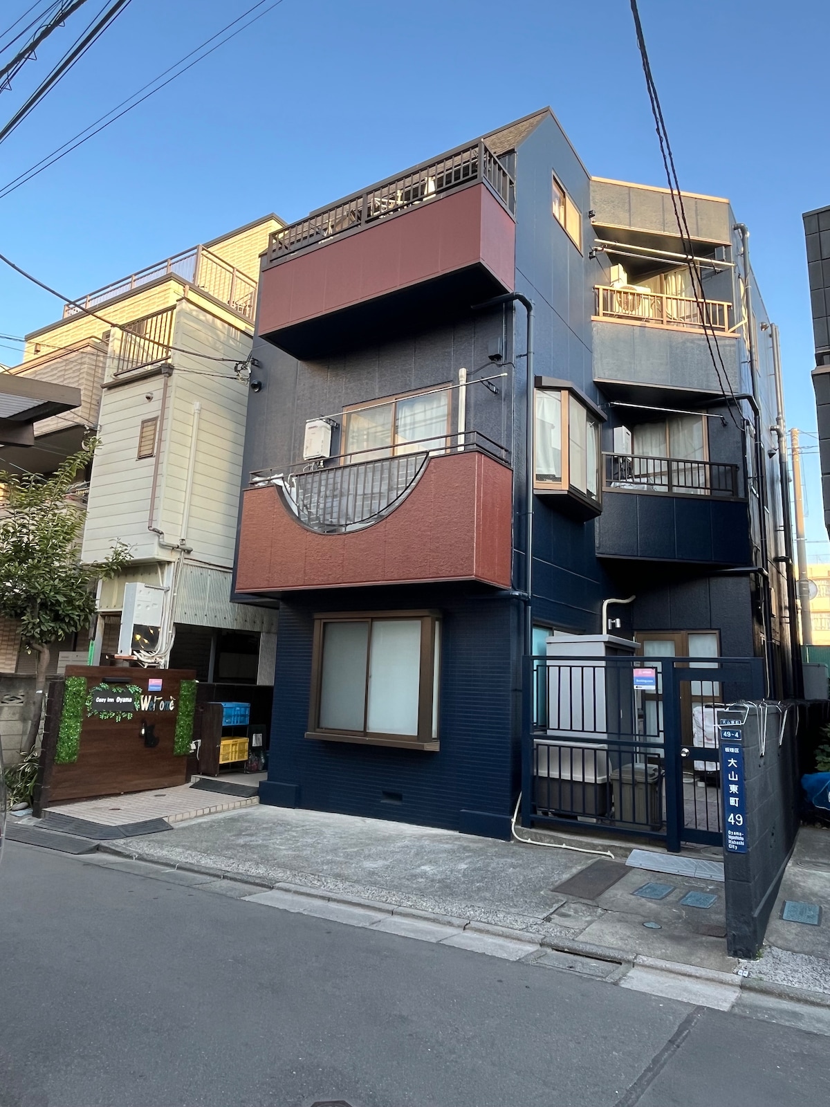 A three-story building is depicted with a mix of dark blue and earthy tones. Balconies are visible on the second and third floors, with windows adorned by light curtains. The surrounding area is characterized by a quiet street and neighboring structures.
