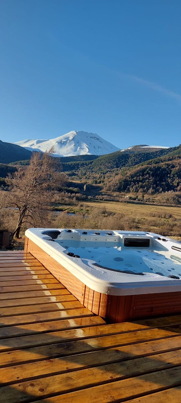 A hot tub is situated on a wooden deck, offering views of the surrounding mountains and valleys. Snow-covered peaks are visible in the background, underscoring the natural beauty of the location.
