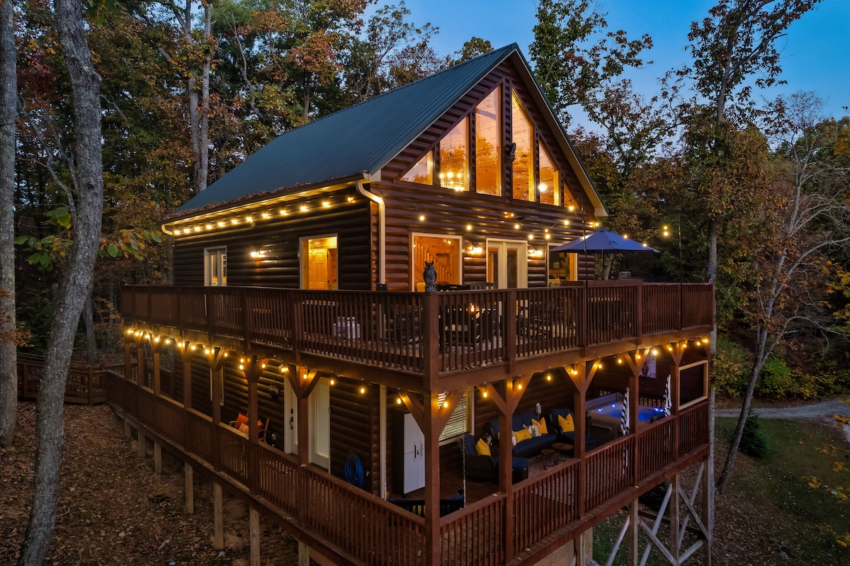 The cabin is set amid a wooded area, showcasing a multi-level structure with a modern design. Soft lighting outlines the decks, enhancing the inviting nighttime ambiance. The spacious wraparound porch features various seating arrangements, offering a comfortable space to relax and enjoy the surrounding nature.