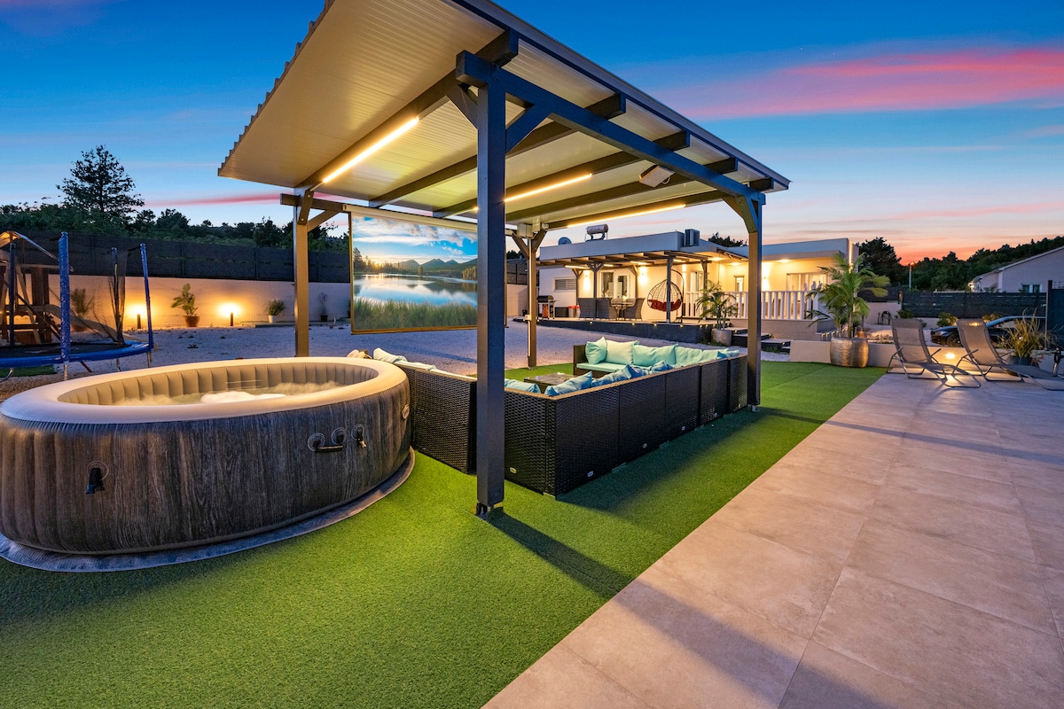 An outdoor area showcases a shaded seating arrangement with fabric cushions, adjacent to a round wooden jacuzzi. A play structure is visible in the background, along with a spacious terrace area designed for relaxation. The setting is complemented by evening lighting, enhancing the tranquil ambiance.