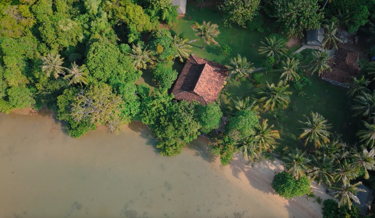 An aerial view of a villa nestled among lush greenery, surrounded by palm trees. The property is situated near a private beach, with clear water gently lapping at the sandy shore. The villa features a traditional design, blending harmoniously with the natural landscape.