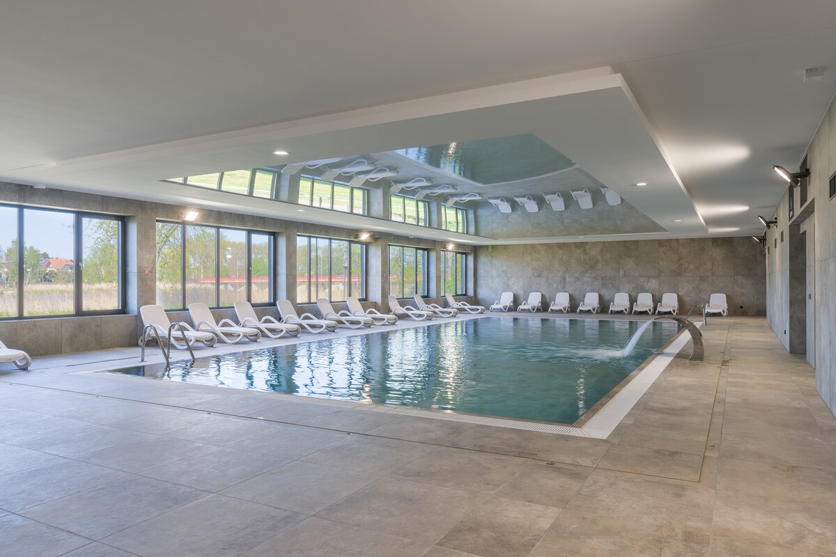 The indoor pool area is surrounded by large windows, allowing natural light to fill the space. Multiple lounge chairs are arranged alongside the pool, creating a relaxed setting. The smooth stone flooring complements the modern design of the facility.