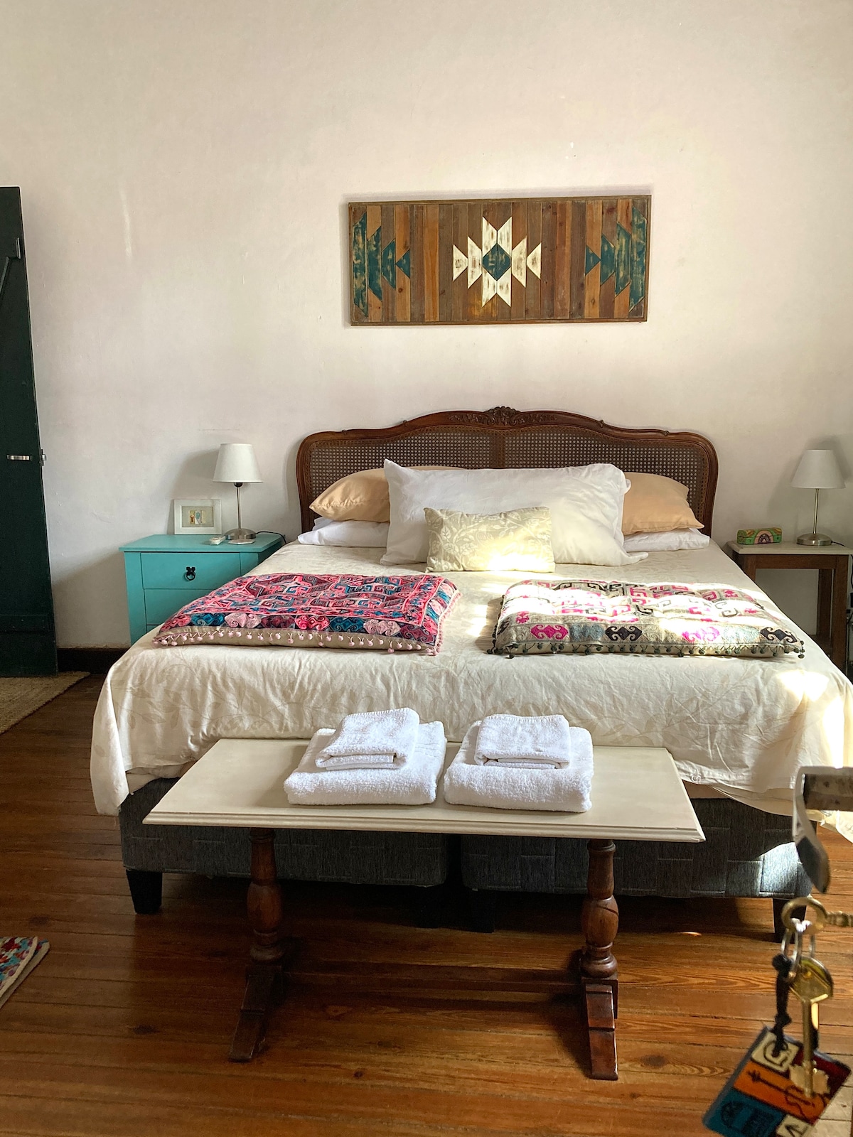 A spacious bed is dressed in cozy blankets and decorative pillows, set against a backdrop of light-colored walls. Two nightstands with lamps flank the bed, while a neatly arranged bench with towels sits at the foot. Light wood flooring adds warmth to the room.