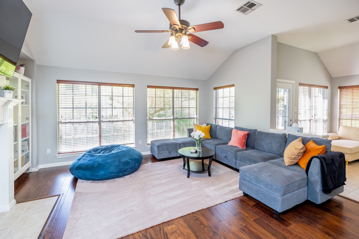 A spacious living room features a large sectional sofa with colorful accent pillows. A circular coffee table sits in the center, and a cozy bean bag rests beside it. Natural light filters through multiple large windows, illuminating the hardwood flooring.