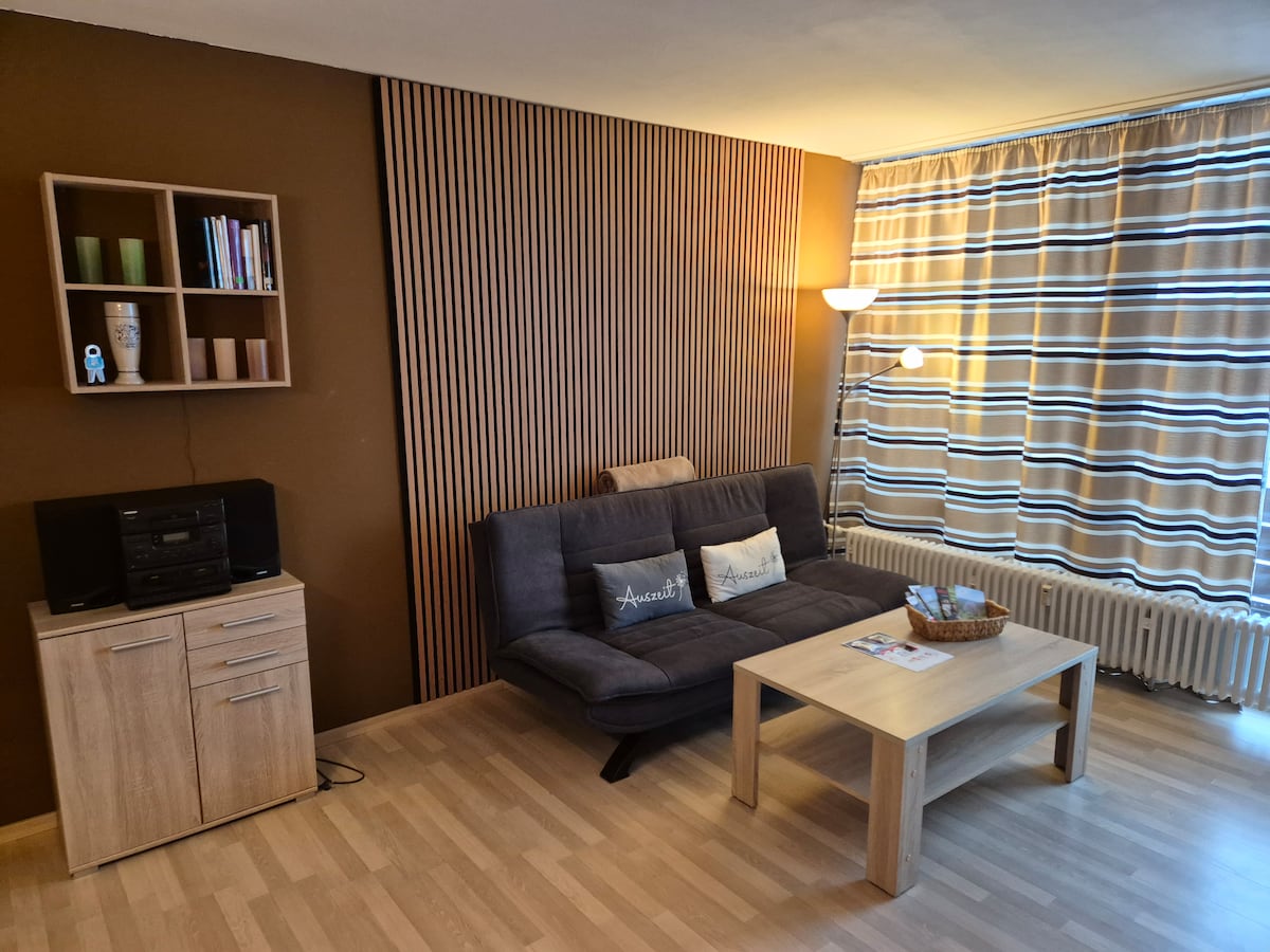 The living area is furnished with a dark gray sofa and a light-toned coffee table. A wooden storage unit holds books and decorative items. Natural light filters through striped curtains, illuminating the space, while a floor lamp provides additional lighting.