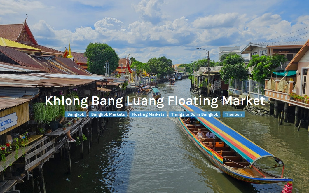 A vibrant scene at Khlong Bang Luang Floating Market features colorful boats navigating the waterway. Wooden structures with traditional architecture line the canal, complemented by lush greenery and clear blue skies. The calm waters reflect the lively atmosphere of local commerce and community activities.