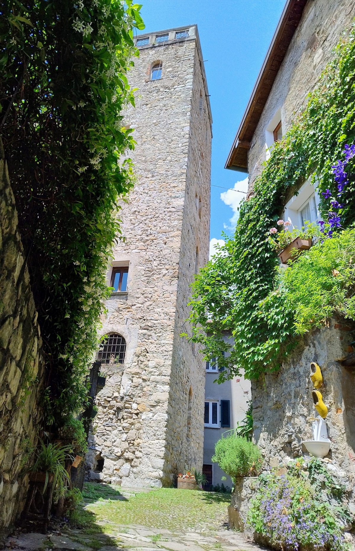 A narrow cobblestone pathway leads towards a tall stone tower, partially covered in greenery. The surrounding buildings reflect historical architecture, with flowers adorning the walls. Bright blue skies provide a backdrop, enhancing the charm of the medieval setting.