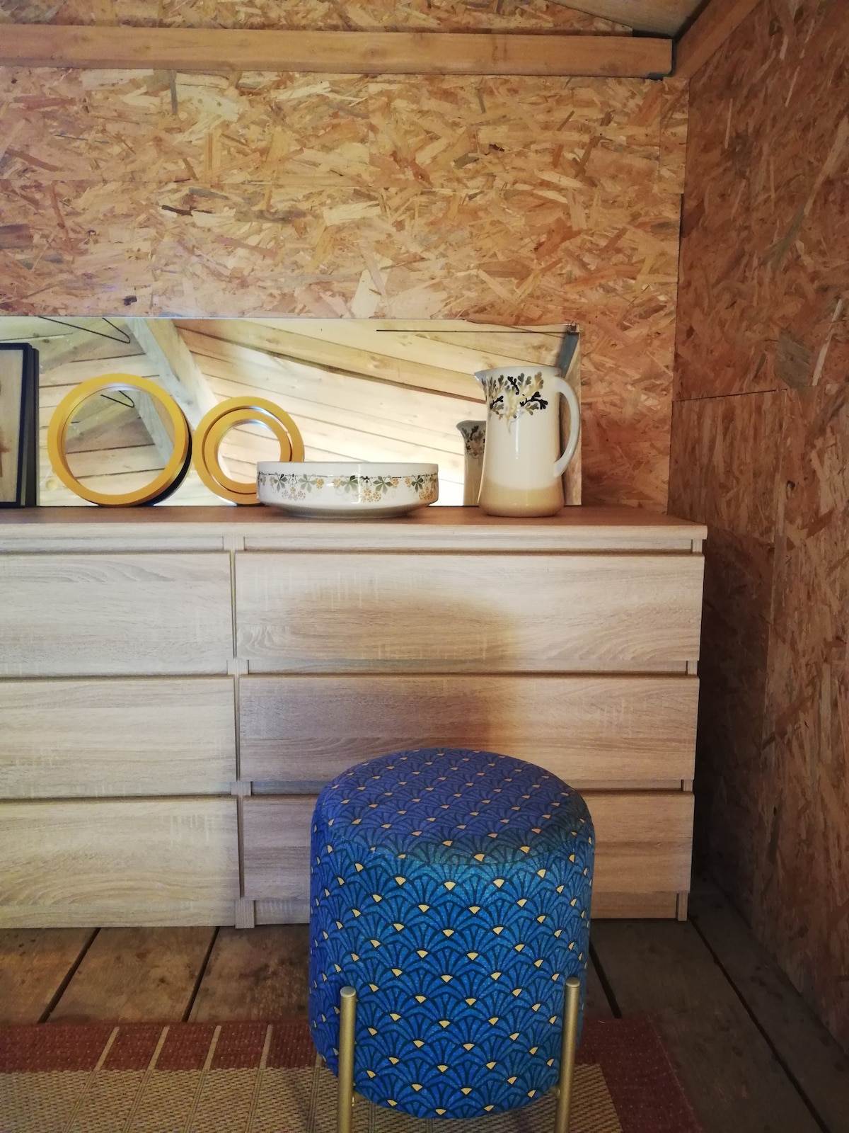 A wooden dresser with multiple drawers is situated against an OSB wall. A decorative bowl and a pitcher are prominently displayed on top, accompanied by circular items in varying sizes. A colorful upholstered stool adds a touch of pattern to the cozy space.
