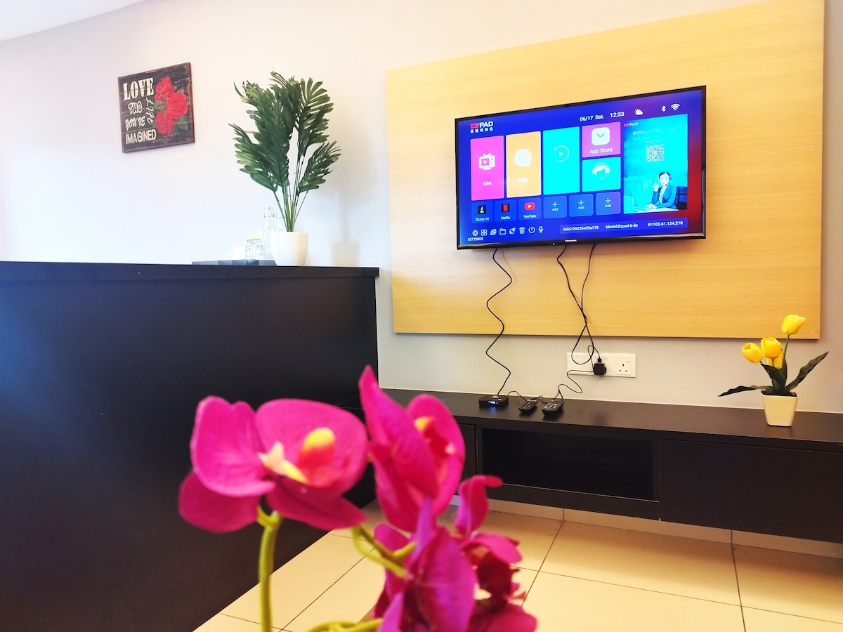 A modern living area showcases a 50-inch television mounted on a light wooden panel, with gaming controllers visible beneath it. A small table features a potted yellow flower, while pink orchids in the foreground add a touch of vibrancy to the space.