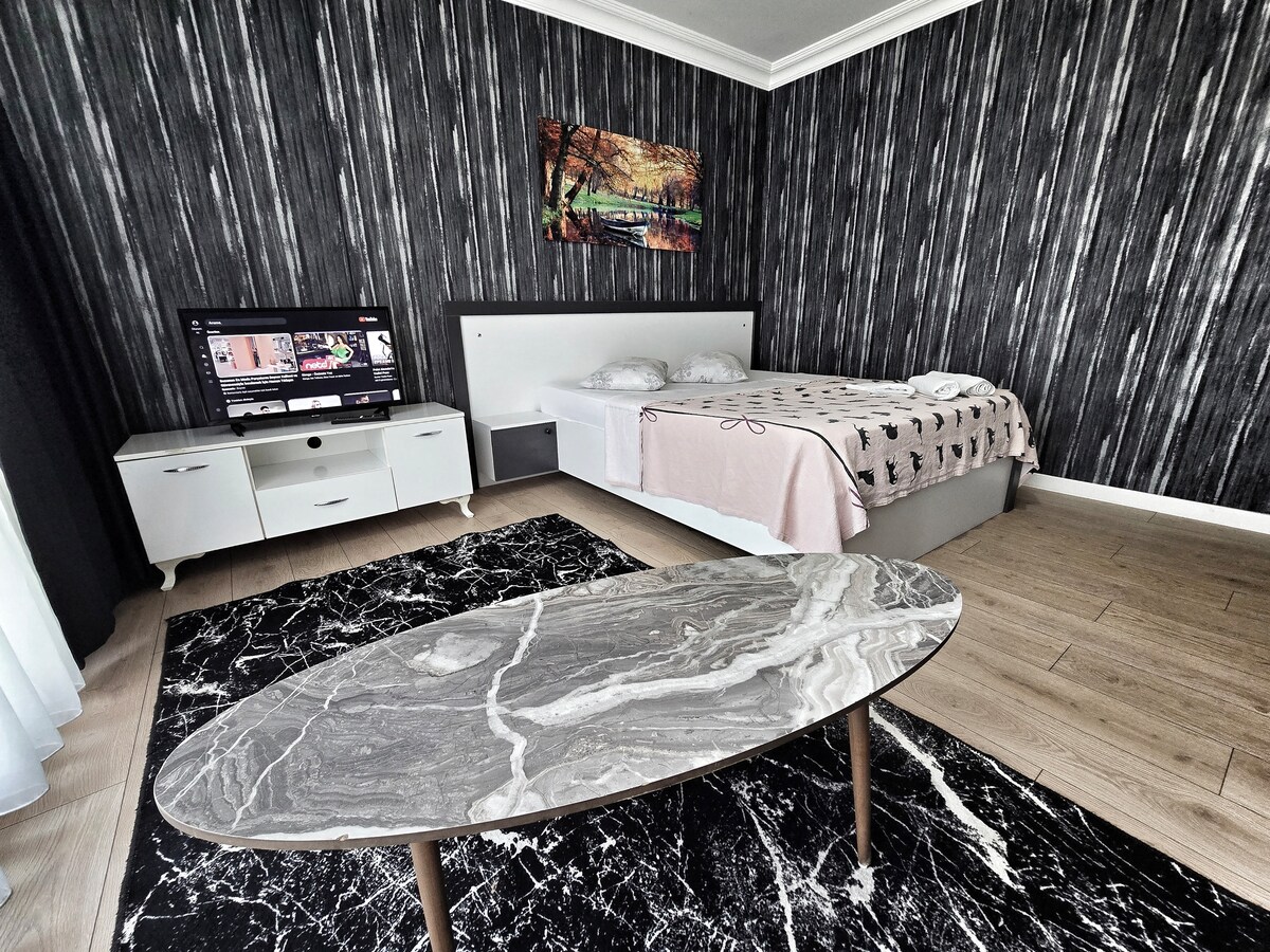 A modern bedroom features a spacious bed with a neutral-toned blanket. A sleek marble coffee table sits centrally on a patterned rug. A flat-screen TV is mounted on the wall, accompanied by minimalistic furniture against the textured black wallpaper.