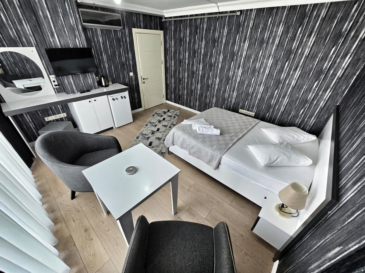 A modern bedroom features a spacious bed with white bedding and two pillows. A small table sits beside an armchair, and a flat-screen TV is mounted on the wall. Textured wallpaper adds depth, while natural light filters through sheer curtains.
