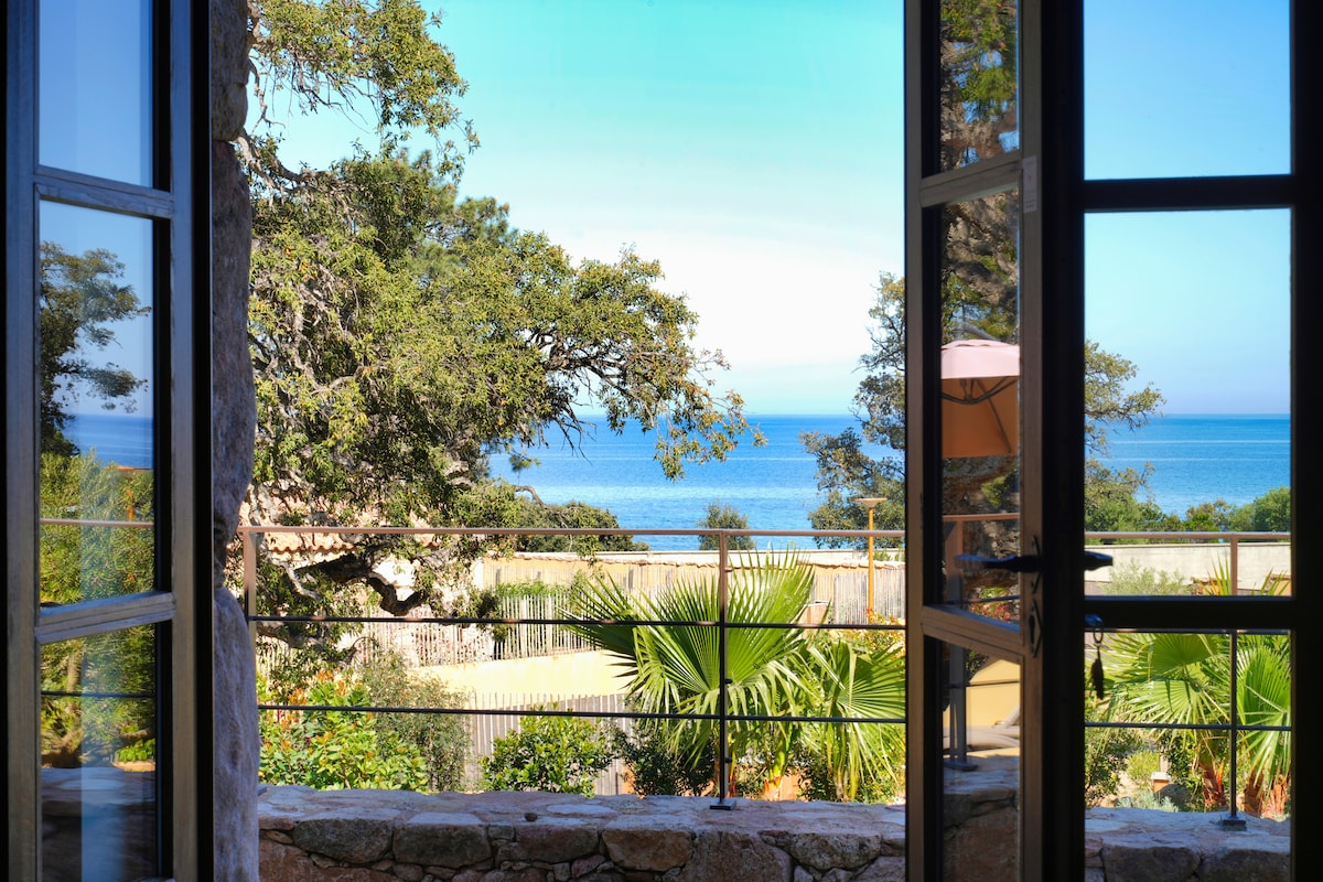 A view of the serene sea and lush greenery is framed by open glass doors. The landscape includes vibrant palm trees and distant coastal waters, creating a peaceful outdoor scene that complements the interior space.
