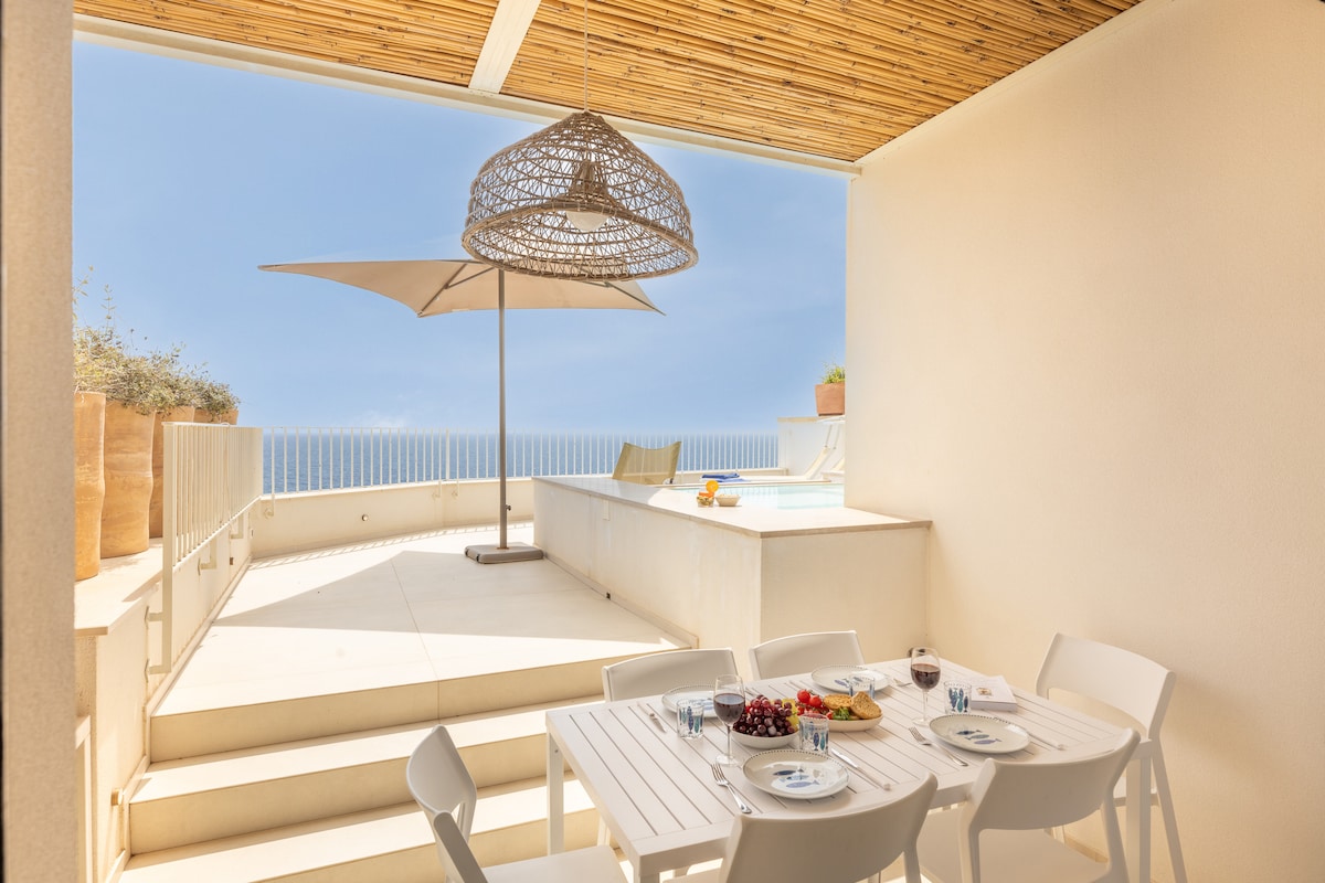 An outdoor dining area features a white table set with dishes and a view of the infinity pool against the sea. A large umbrella provides shade, while potted plants add a touch of nature. Steps lead down to the pool area, enhancing the seamless connection to the landscape.