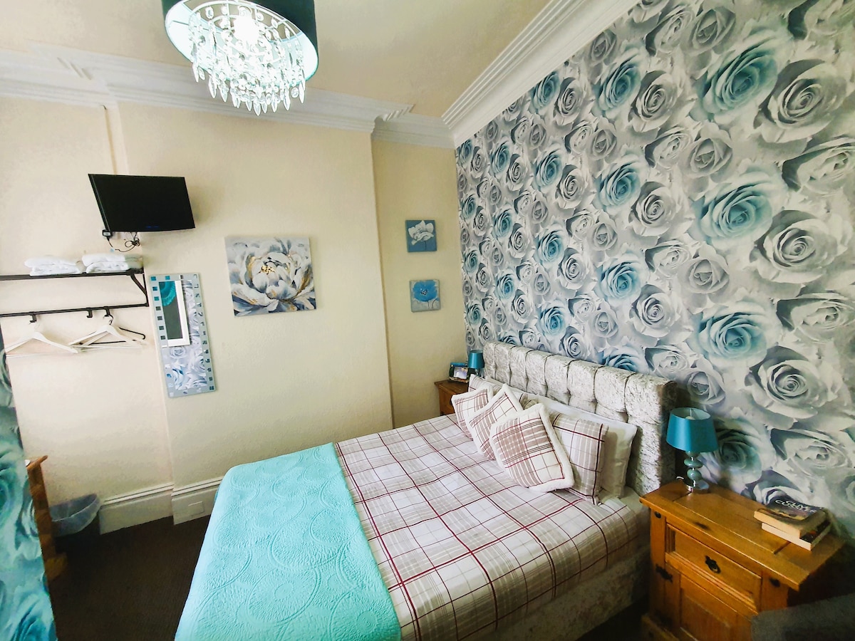 A guest room is featured with a spacious bed adorned with light blue and white patterned linens. The walls showcase a floral design in shades of blue and white. A flat-screen TV is mounted nearby, and bedside tables provide additional convenience.