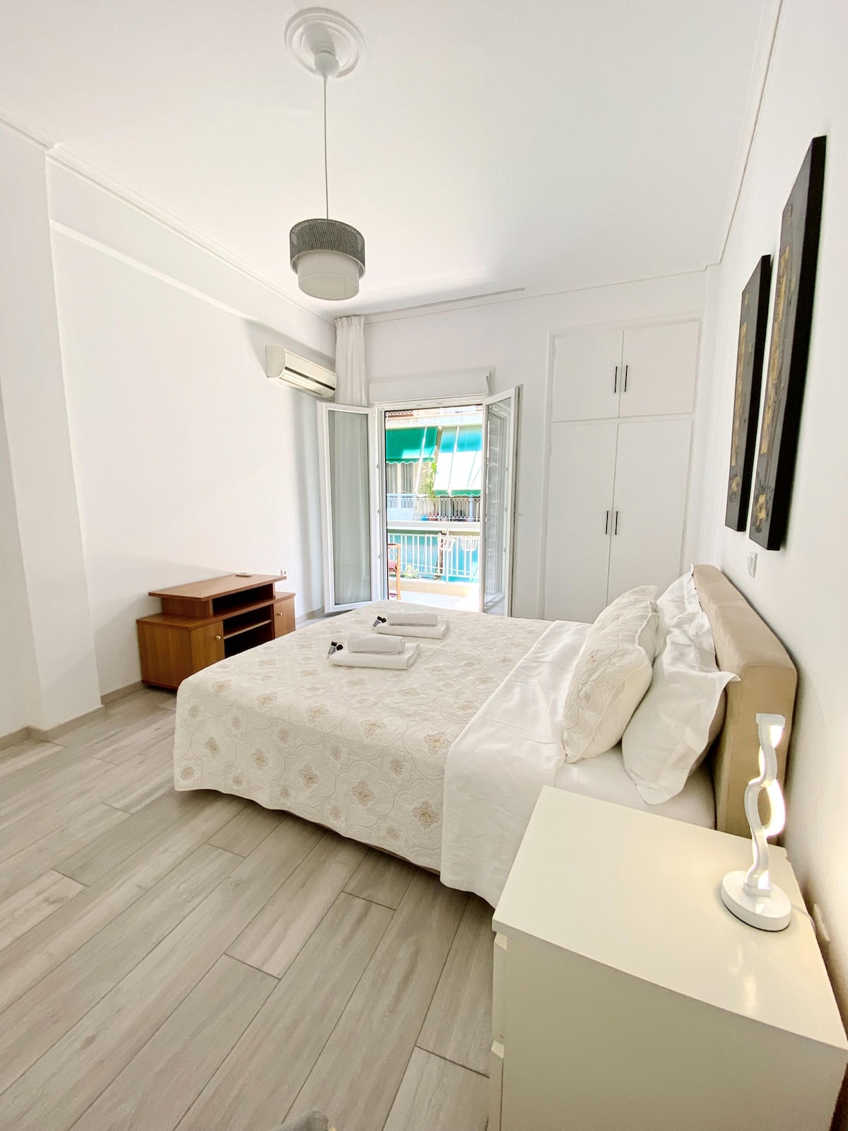 A light and airy bedroom features a spacious bed with neutral bedding and decorative pillows. A wooden dresser is positioned against one wall, while a closet provides additional storage. Natural light filters in through an open balcony door, enhancing the serene atmosphere.