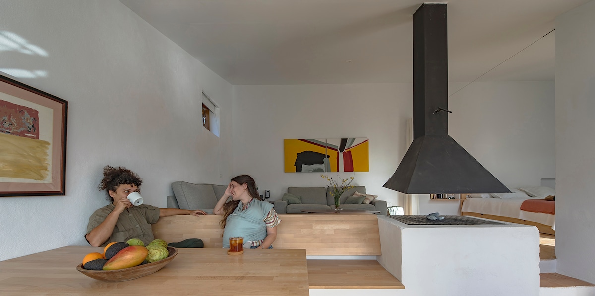 A spacious open-concept living area features a wooden dining table with a variety of fresh fruits. A modern chimney stands prominently, serving as a focal point. Comfortable sofas are arranged nearby, and colorful wall art adds visual interest to the bright, welcoming space.