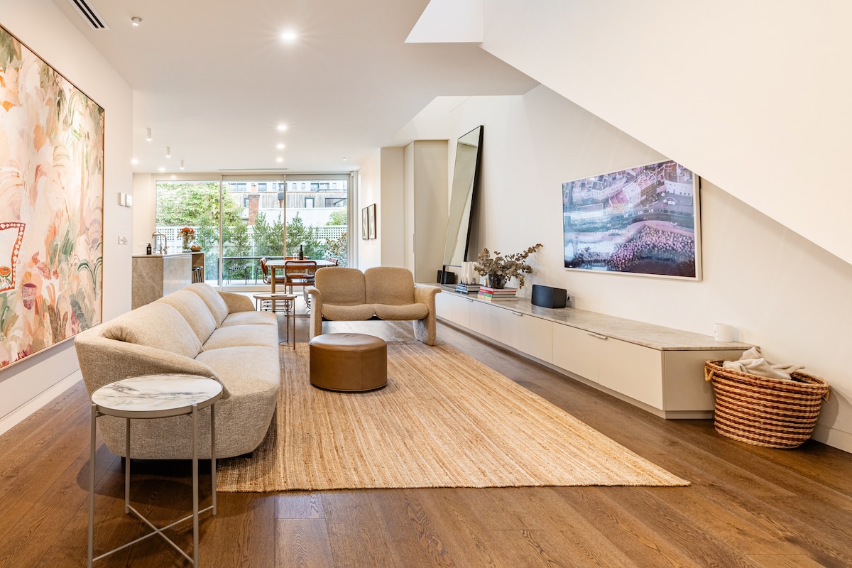 A spacious living area is designed with modern furnishings, featuring a light-colored sofa and two accent chairs. A neutral area rug grounds the seating arrangement, while large windows provide natural light and views of greenery outside. A simple wall-mounted television adds to the contemporary decor.