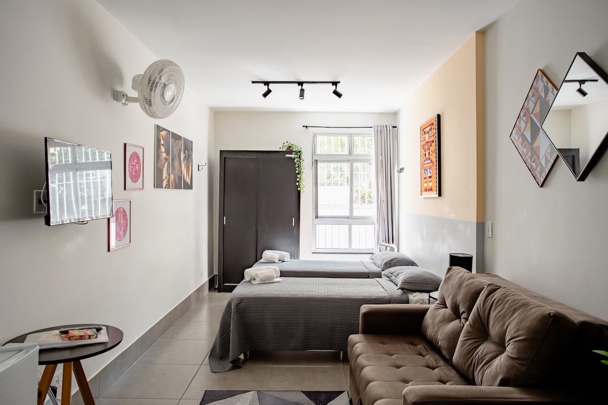 A spacious studio apartment is featured with a comfortable sofa and a bed neatly made with soft bedding. A flat-screen TV is mounted on the wall, alongside decorative wall art. Natural light filters in through a large window, enhancing the airy feel of the space.
