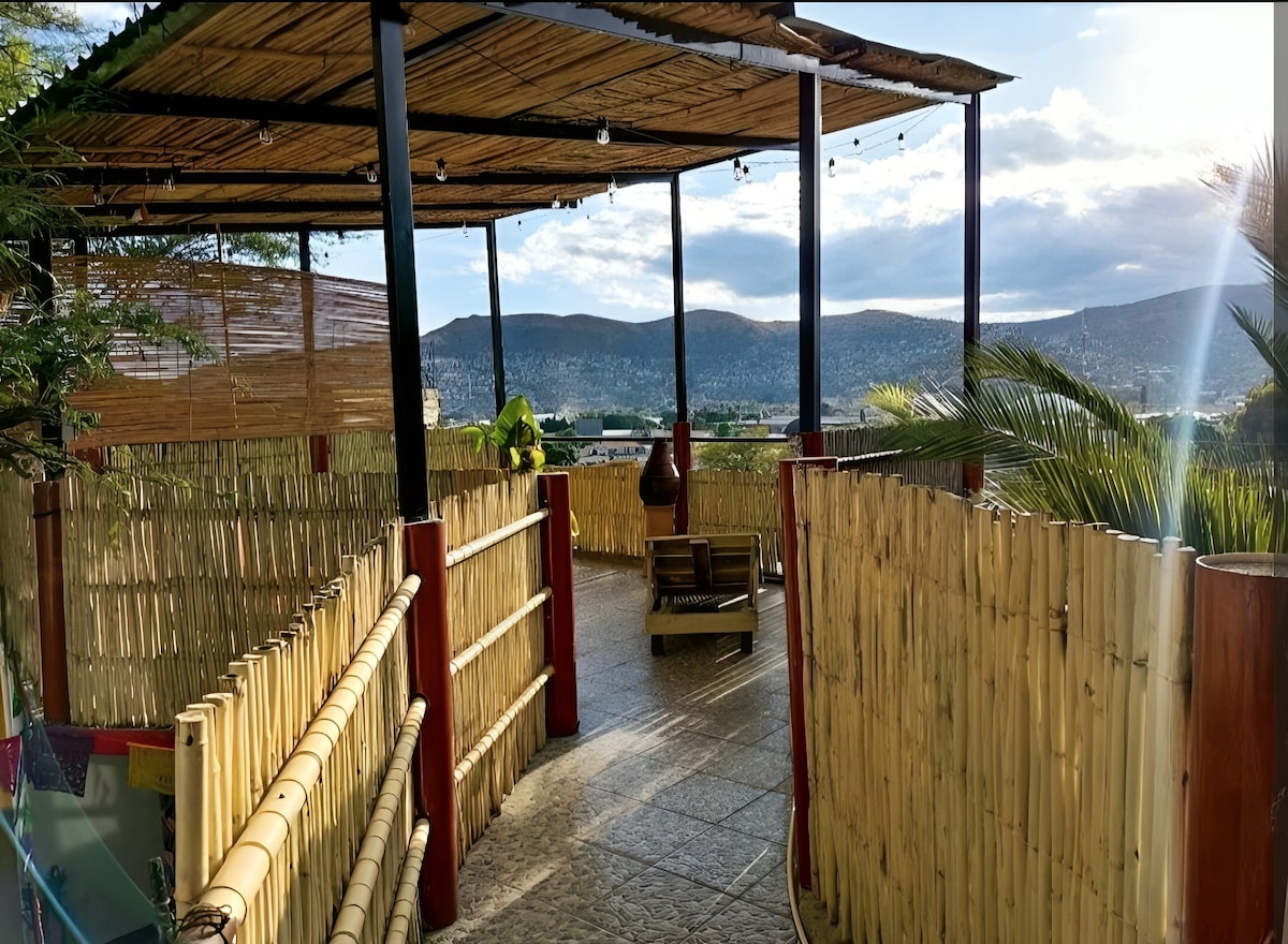 An open terrace is framed by bamboo fencing, featuring a paved floor and a wooden seating area. The space offers panoramic views of the surrounding hills, with warm sunlight streaming through the clouds, creating a serene outdoor environment.
