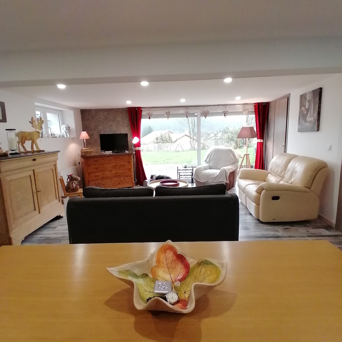 A spacious living area is visible, featuring soft seating arrangements and a wooden television console. Large windows are dressed with red curtains, allowing natural light to illuminate the interior. A decorative bowl with vibrant leaf motifs sits on a wooden table, adding a subtle touch of color.