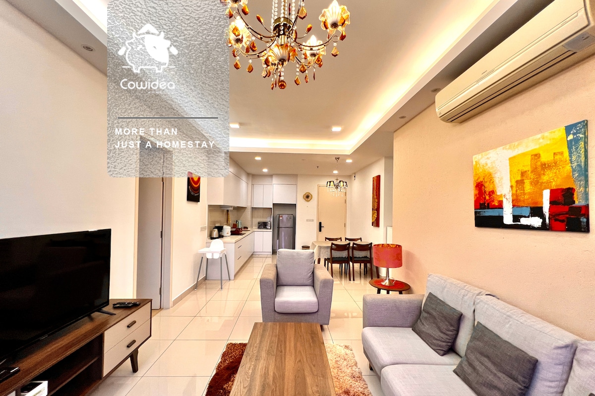An elegantly furnished living space is shown, featuring a soft grey sofa facing a modern television. A dining area is positioned in the background with a round table surrounded by four chairs, while a bright chandelier hangs above. Colorful artwork adds a vibrant touch to the walls.
