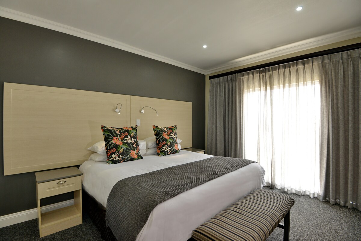 A spacious bedroom is featured, with a large bed adorned with decorative pillows. Soft lighting complements the elegant decor, while large windows, dressed with sheer curtains, allow natural light to fill the room. A bench sits at the foot of the bed, providing additional seating.